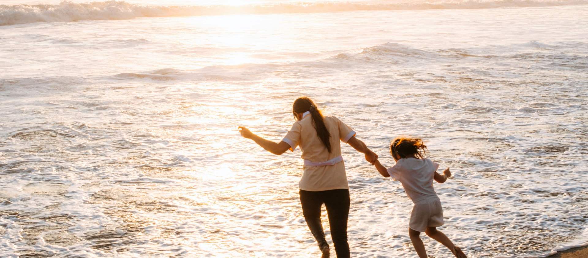 A mother and daughter holding hands, jumping over the waves at sunset