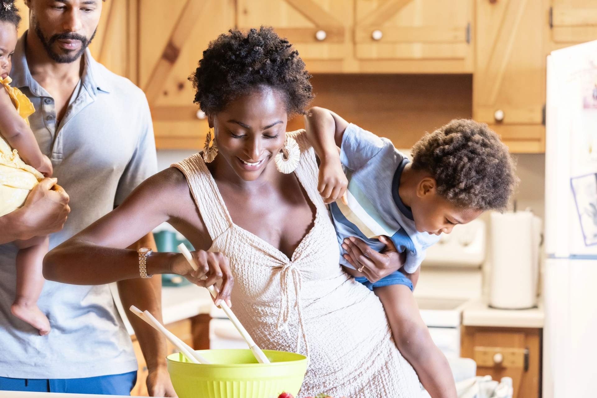 A smiling mother mixing a salad while holding her son, with the father and baby beside her