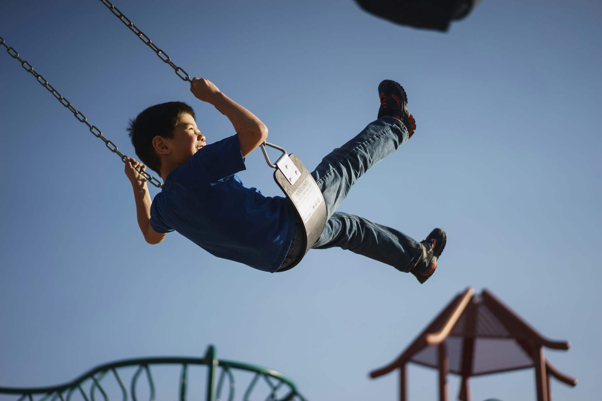 A happy boy swinging high on a playground swing under a blue sky